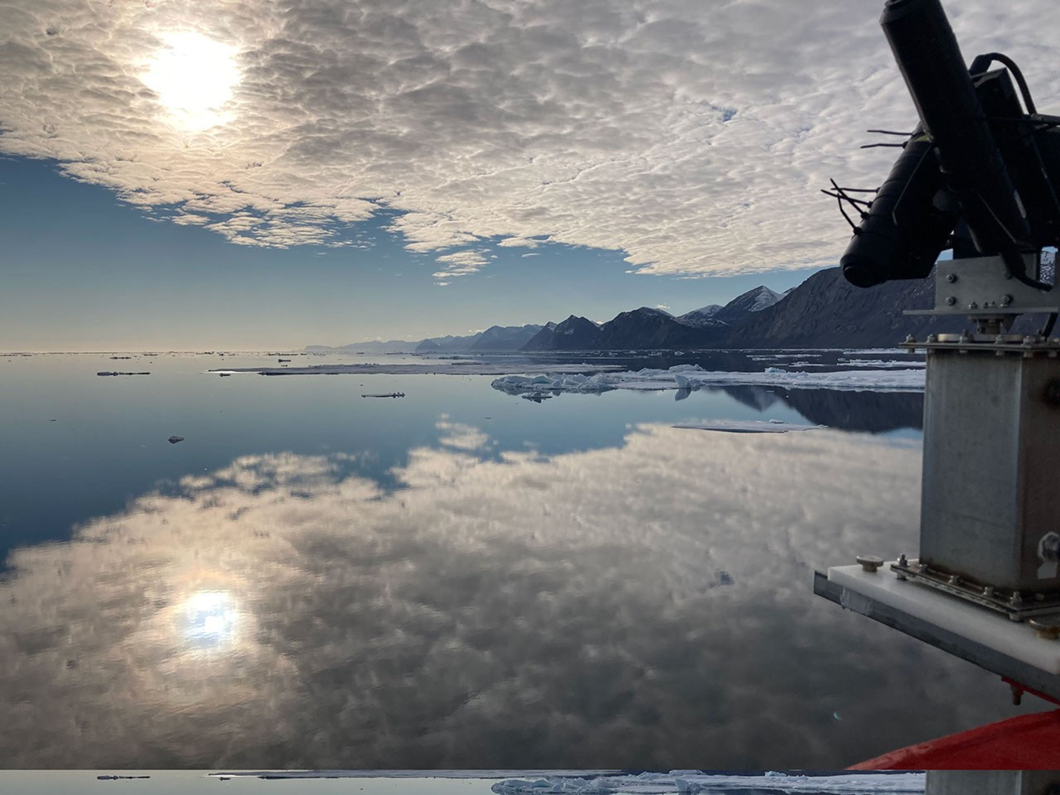 An instrument with radiometers is shown mounted on the bow of a research ship in one corner of a glorious view of an oceanic channel so still that the sky is reflected in a mirror image. Through a layer of cloud, the sun has rainbow edges.
