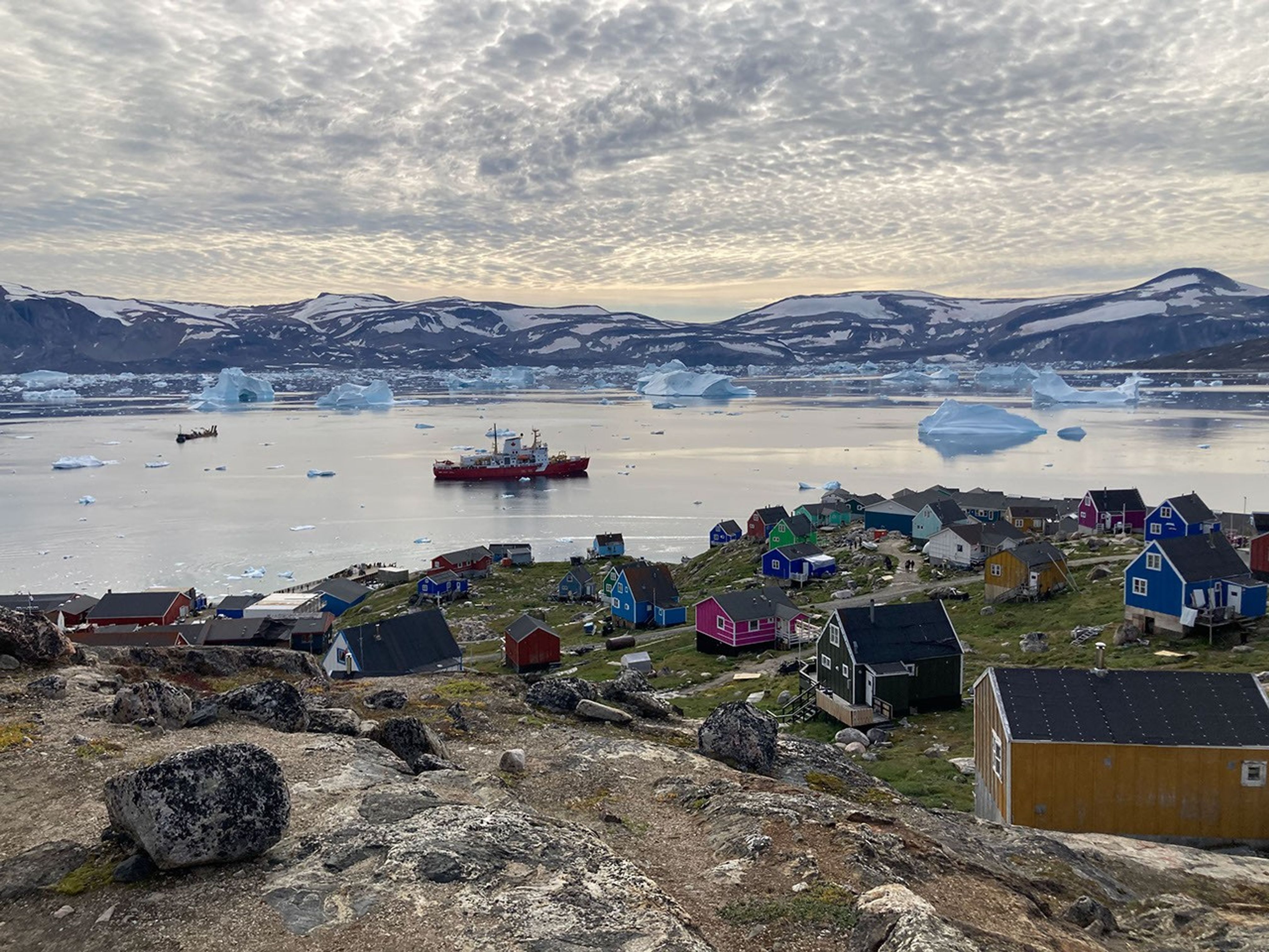 A glowing cloudy sky casts a pearly light over a bay dotted with icebergs. In the foreground, small boxy houses perch on a grassy hillside speckled with patches of snow.