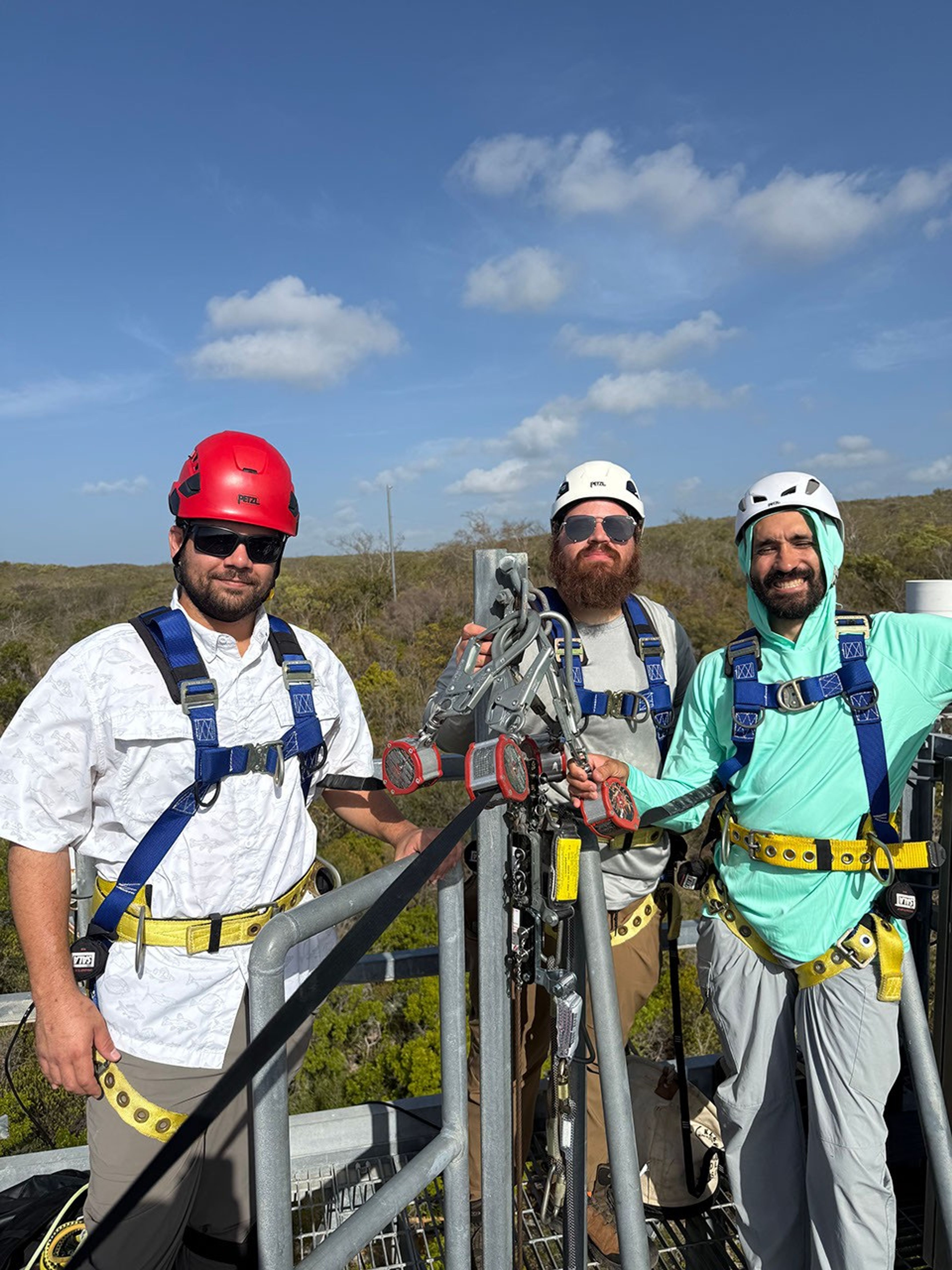 Three men in hard hats, outdoor gear, sunglasses, and safety harnesses stand atop a metal observation tower.  