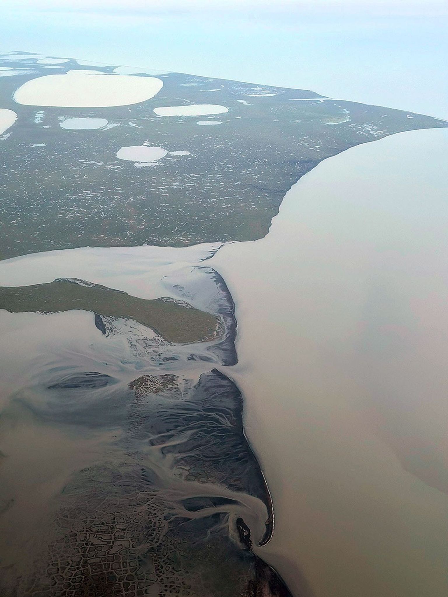 An aerial view of dark grey-green coastline, dotted with ponds, juts into pale blue, open sea. Cloudy rivers flow toward the coast.