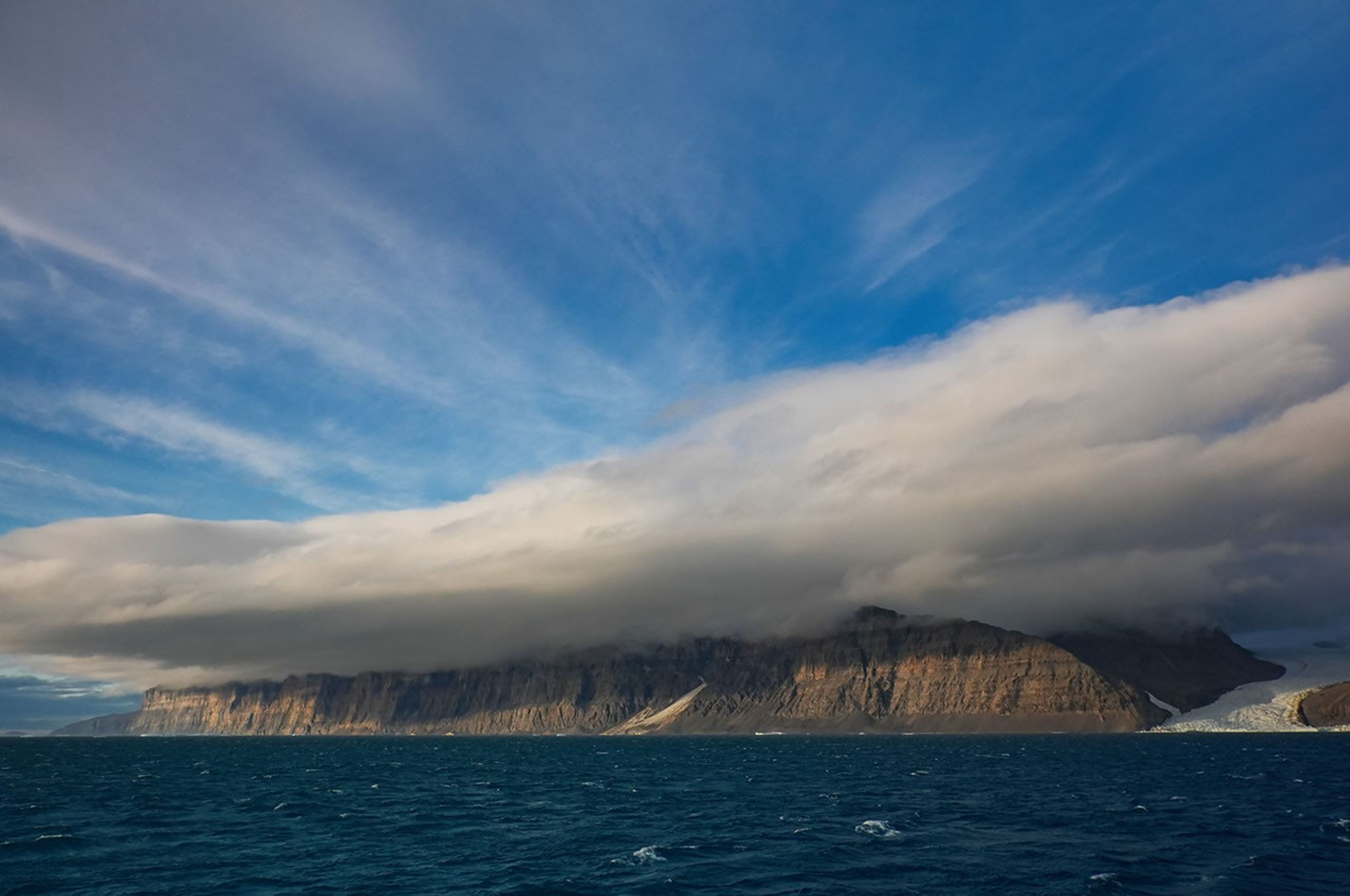 A brown rocky cliff emerges from a dark blue sea. It is capped and nearly overwhelmed by a low-hanging cloud.