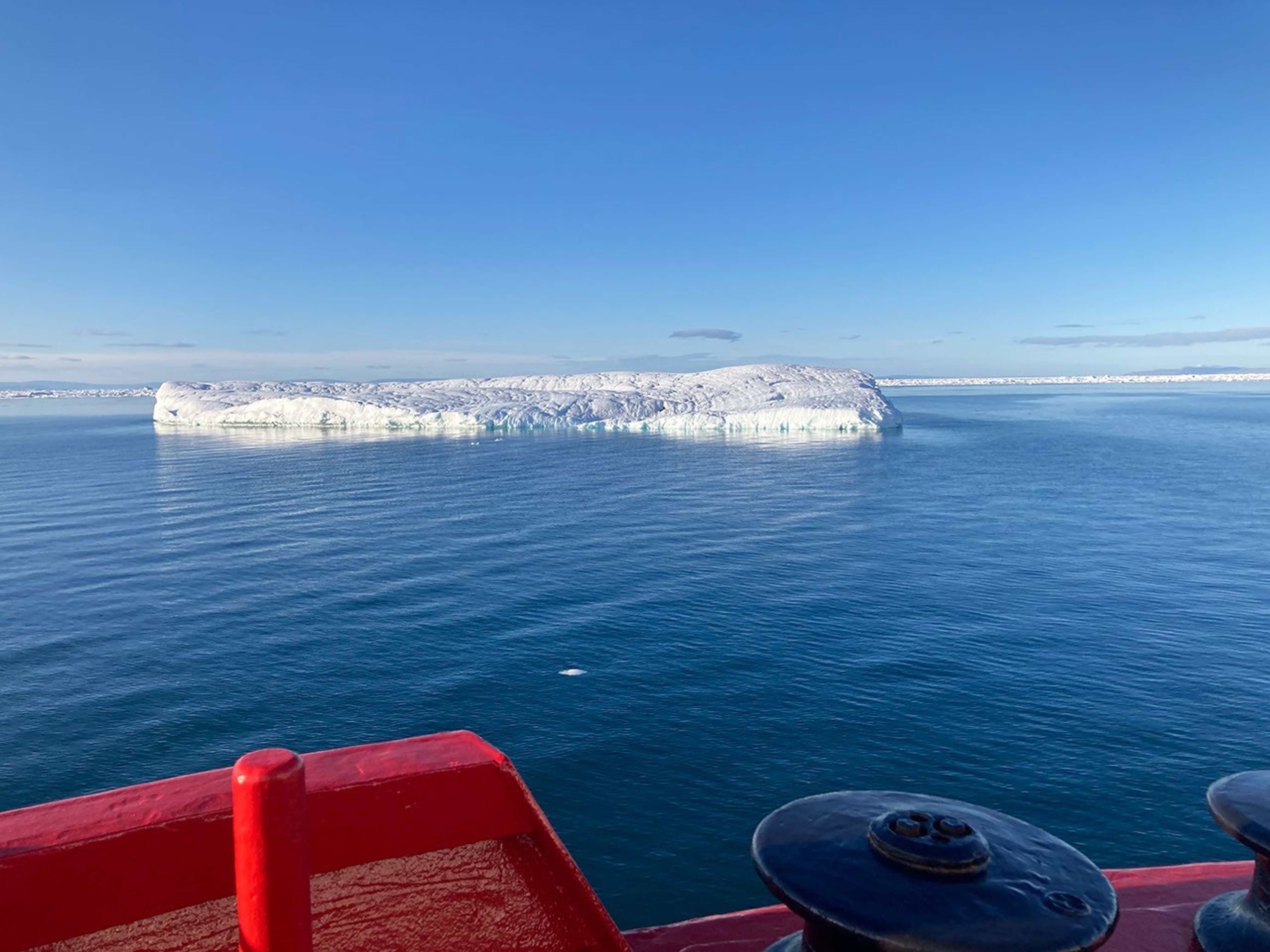 A low, nearly flat ice berg floats not far from the research ship’s red rail.  
