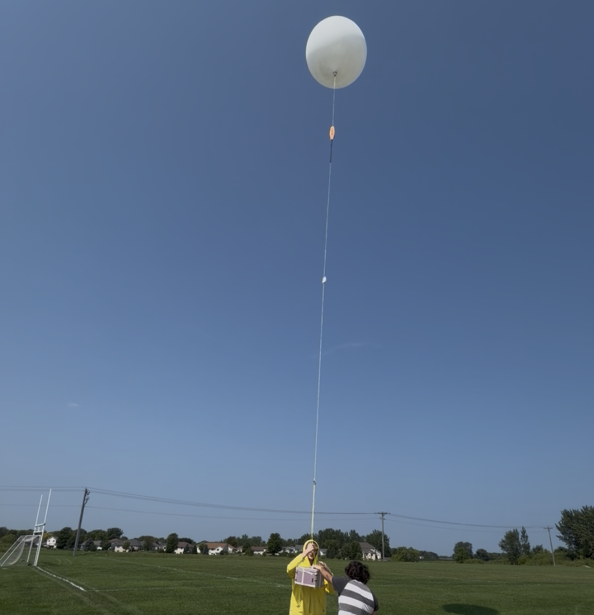 A weather balloon reaches the height of its tether aboard its launcher, a student clad in yellow, and another helper.
