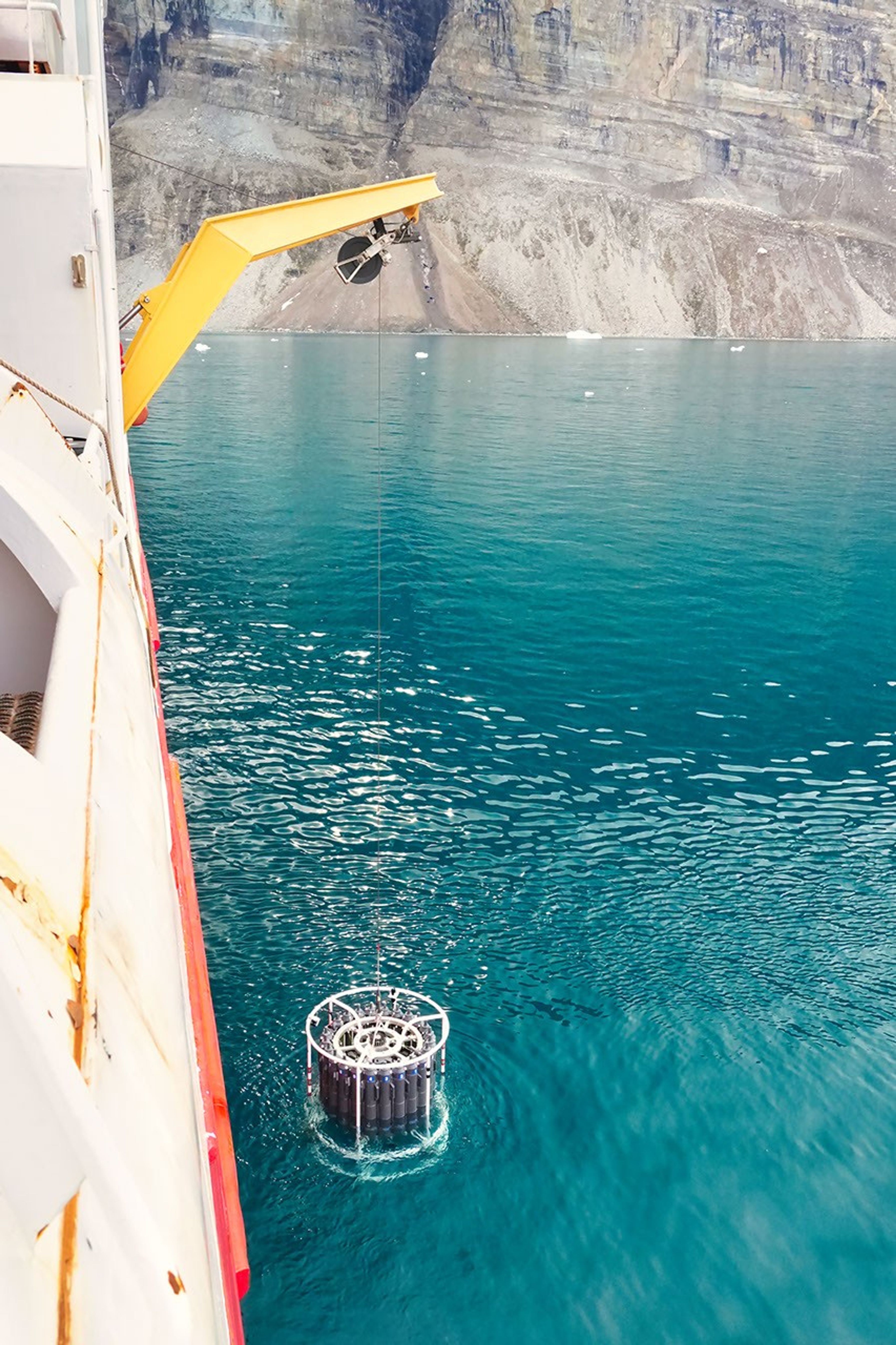 The ship’s yellow crane extends over a turquoise sea; its cable descends into the sea. The gray, tan, and white striations of the glacier front’s ice tower in the background.