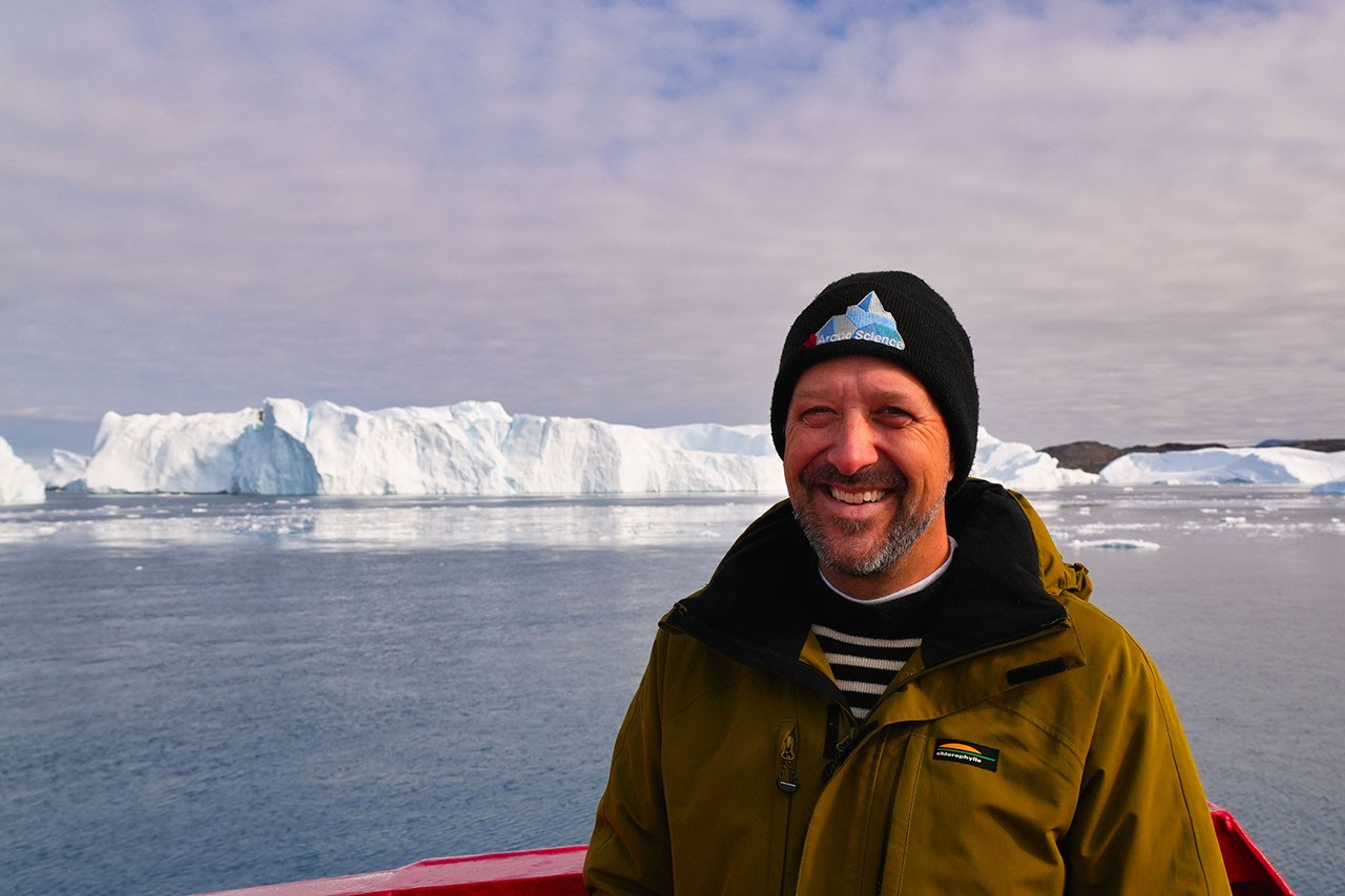 A scientist in outdoor garb poses in front of open water that leads to the white ice cliffs of a glacier front.  