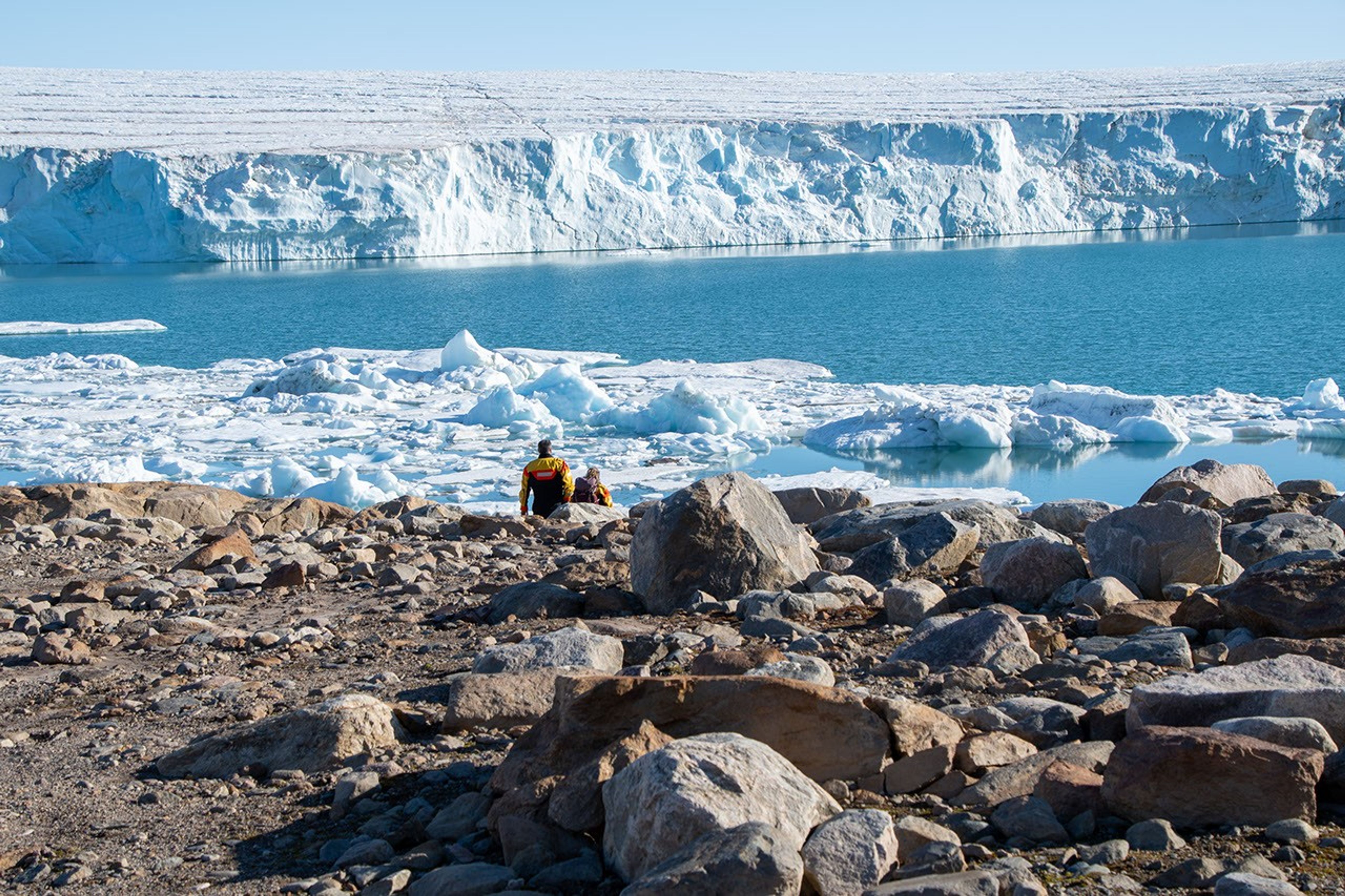 Two people in outdoor clothing explore a rocky shore. In the background : sea ice, open water, and the white ice cliffs of a glacier front.  