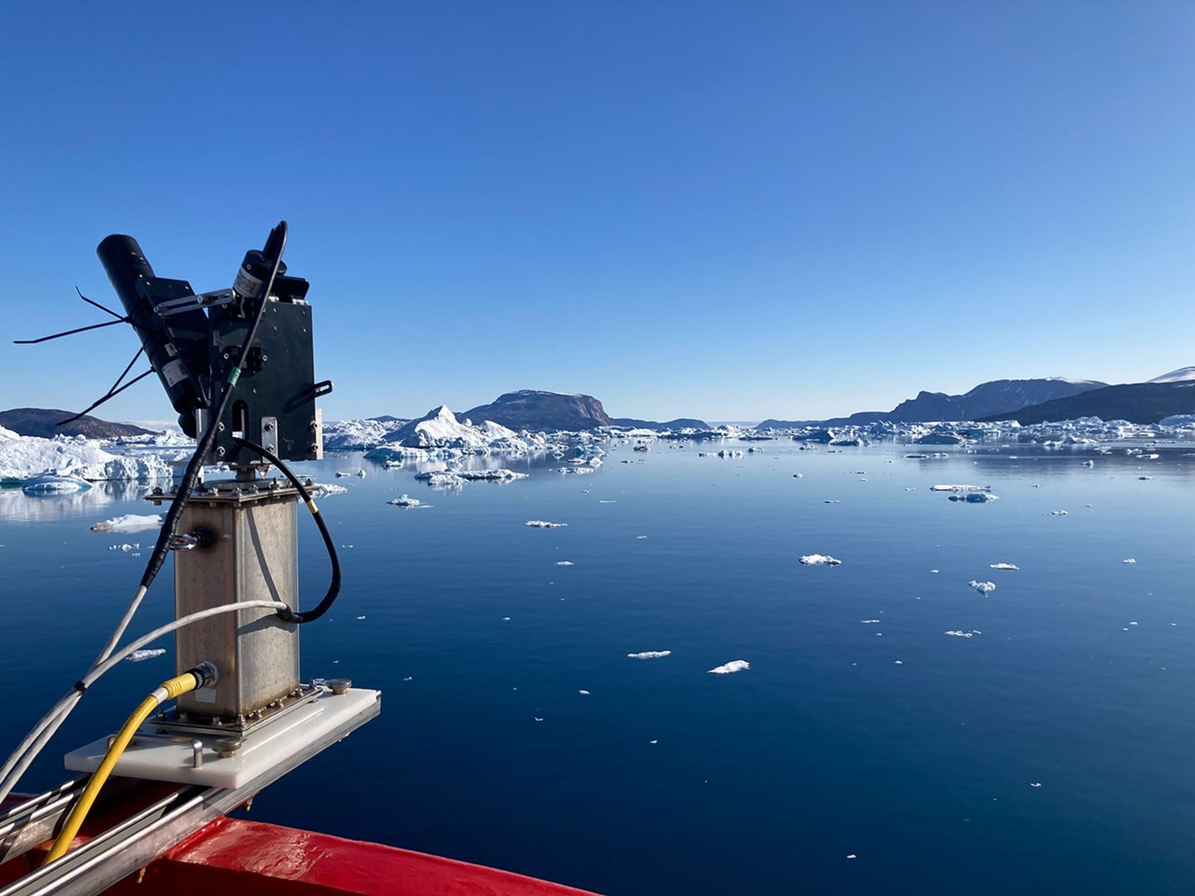 An instrument with radiometers is mounted on a gray metal « tower » aboard the ship. It overlooks a placid deep blue sea edged with icebergs and purple hills.  