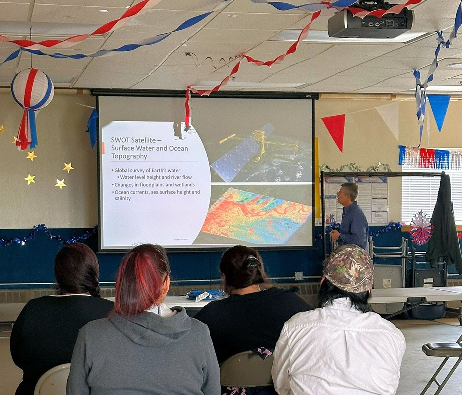 A man makes a slide show presentation in a community hall. Our perspective is from behind several rows of folding chairs, and several audience members. They include a toddler who stands between her parents, facing the camera.