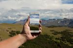 A hand holds a cell phone against a background of rolling hills and a cloudy sky. The cell phone shows an image of the landscape in the background.