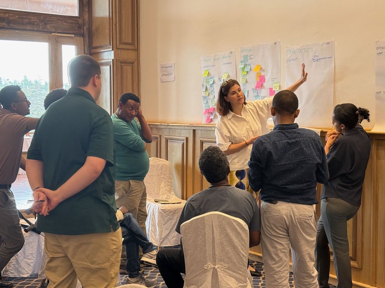 A person stands at the wall with paper pads on it and sticky notes on those. A small crowd engaged participation with the speaker are mostly standing around and seemingly deep in thought.