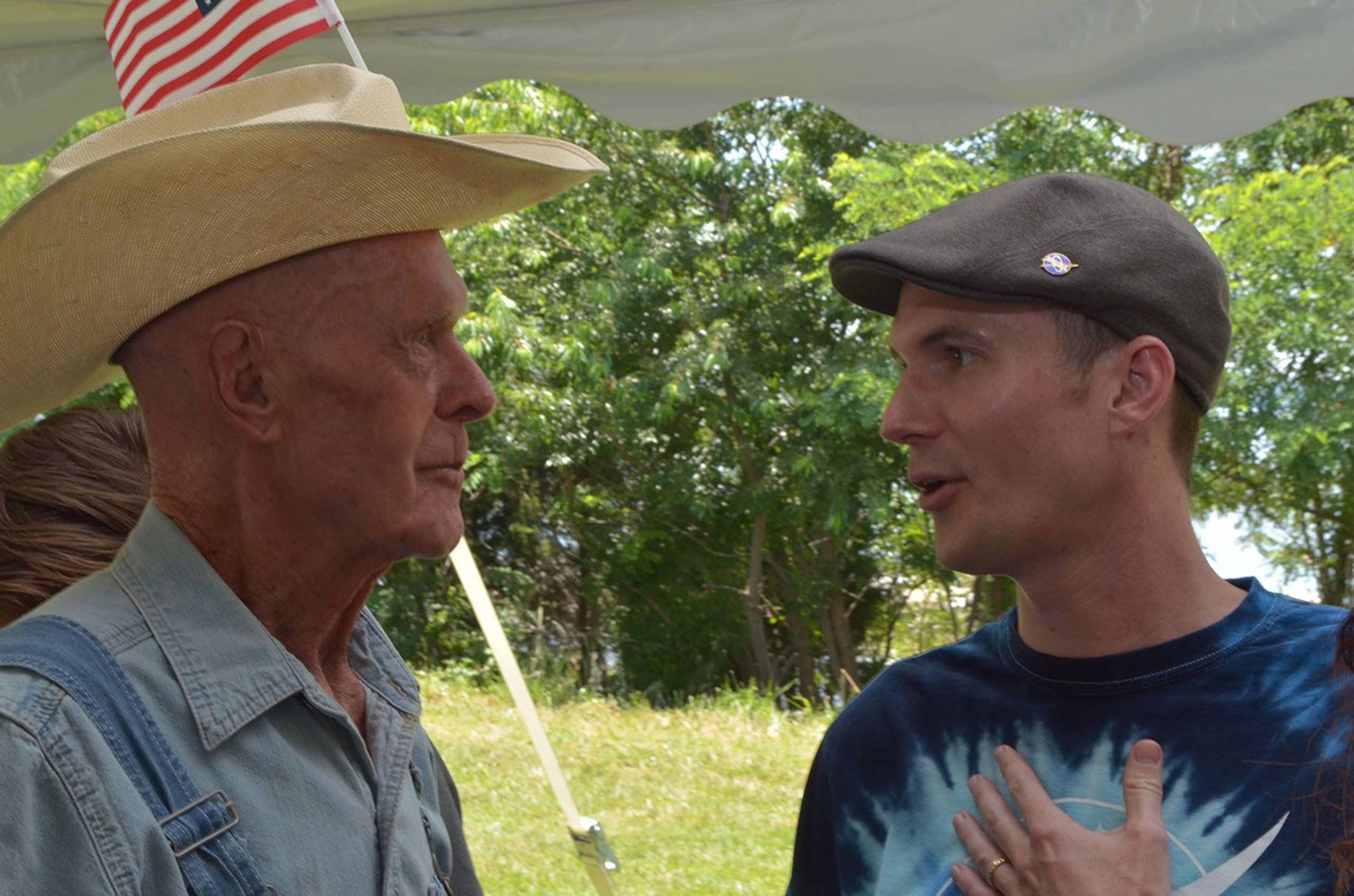 elder farmer on left in a straw hat talks to a man in a beret and tie-dye shirt in front of a nearby woodline