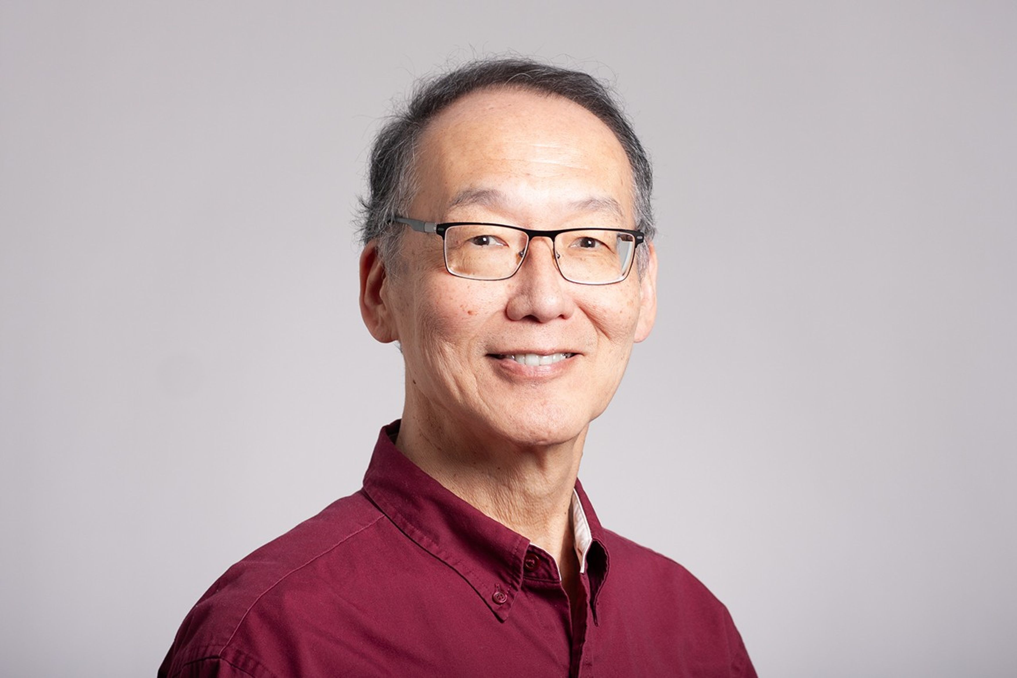 Asian man in a red button down shirt in front of an neutral gray-white background.