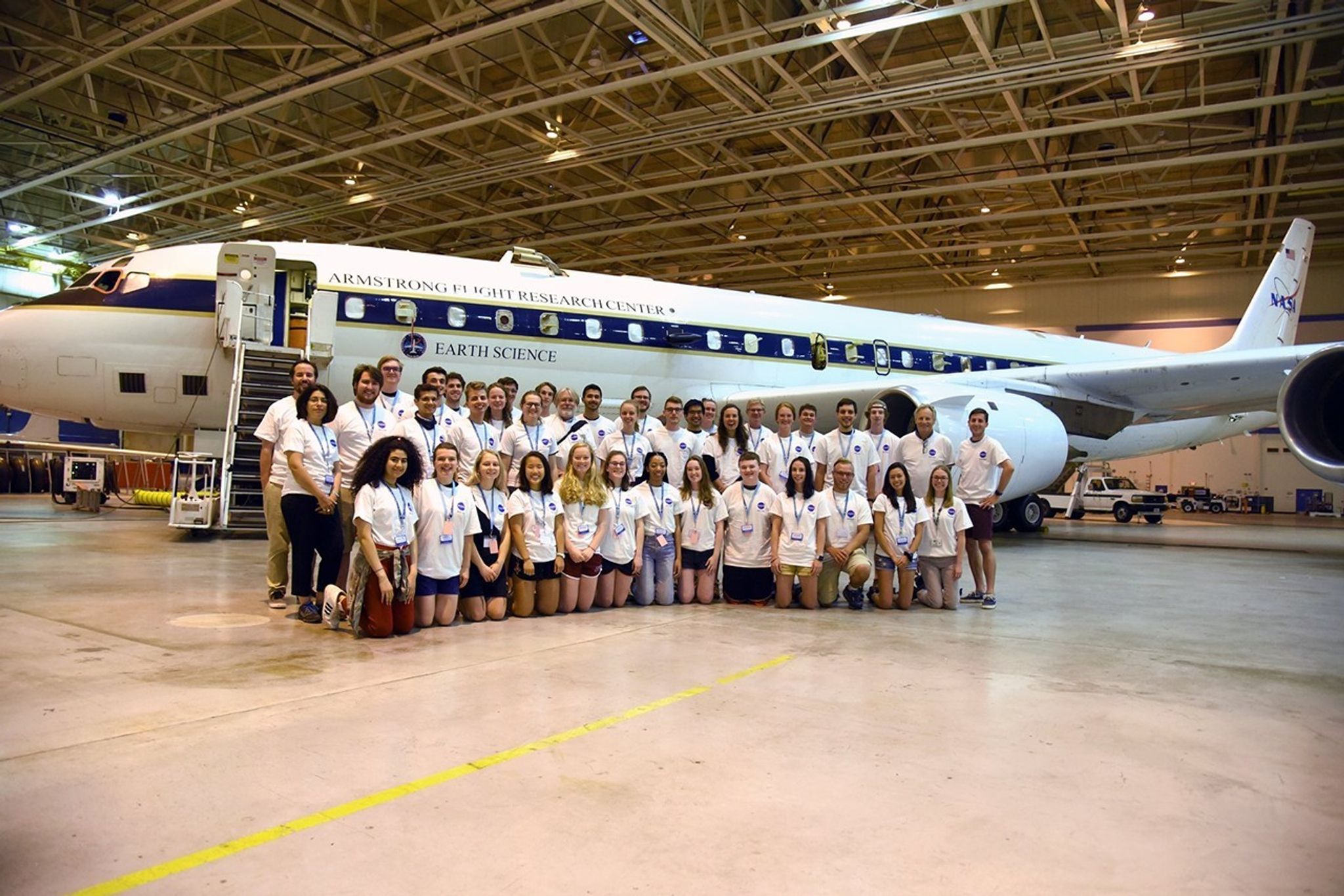 Two rows of students, all in white T-shirts, pose in front of a plane branded with the NASA logo and Earth Science, inside a hangar.