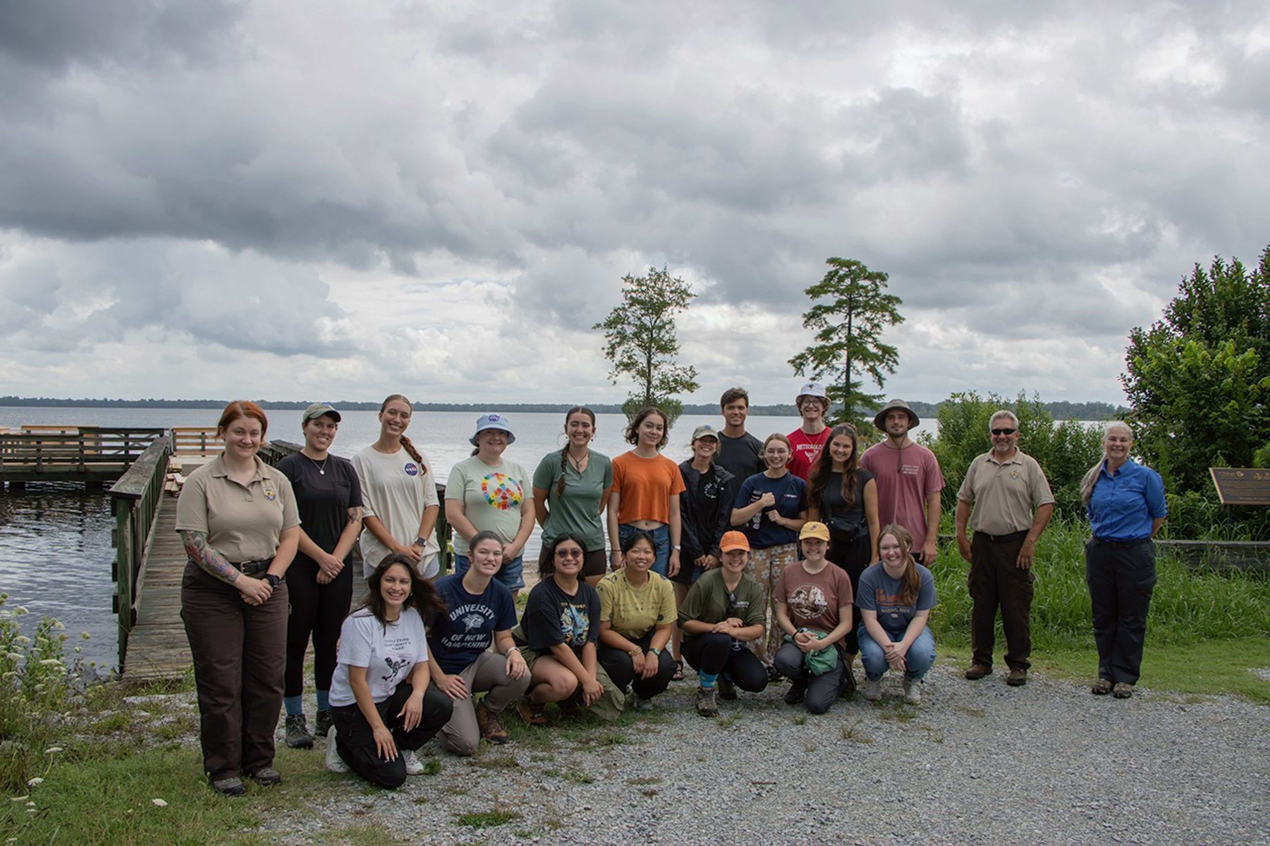 A group of students stand on a gravel path in front of a dock that extends into a body of water with treelines visible on the other side on a cloudy day.
