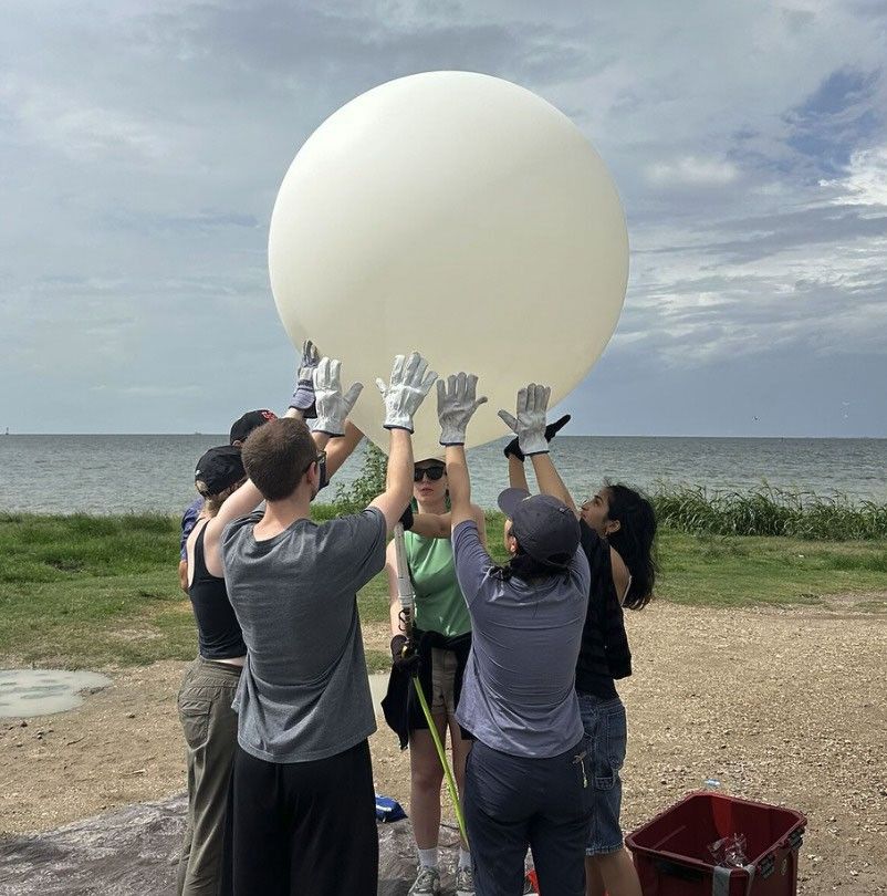 Five people, from SARP Atmospheres 2026 cohort, stand on a shoreline with a body of water and cloudy sky behind them. They are gathered in a circle, wearing thick work gloves, and reaching their hands up to support a large white weather balloon as they prepare to release it.