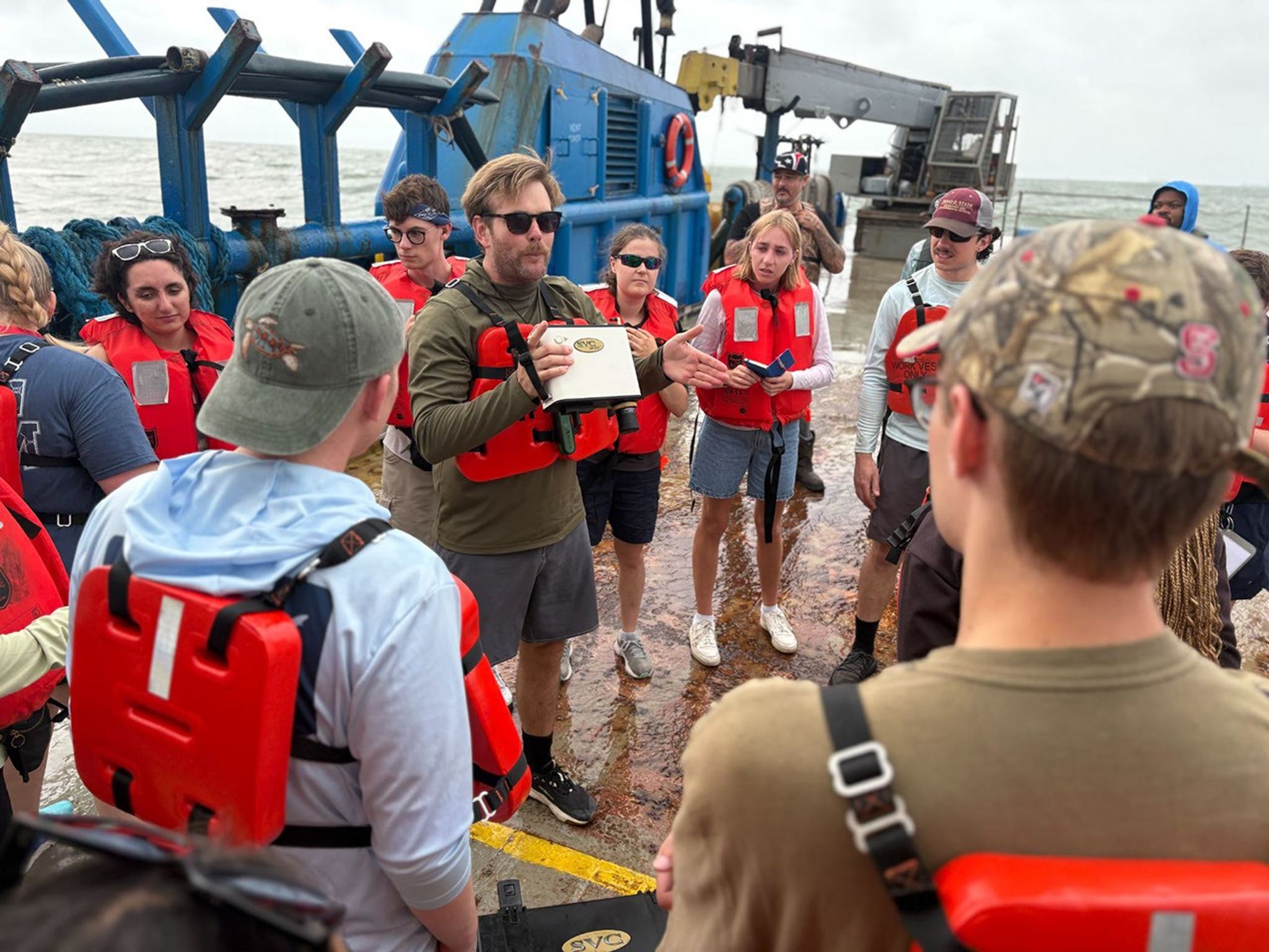 A group of people from SARP Oceans 2026 wearing orange life jackets are gathered in a semicircle on the deck of a research vessel. In the center, a man, Henry Houskeeper, holding a small, box-like piece of equipment gestures as he explains it to the group. Large blue marine equipment and the ocean are visible in the background.