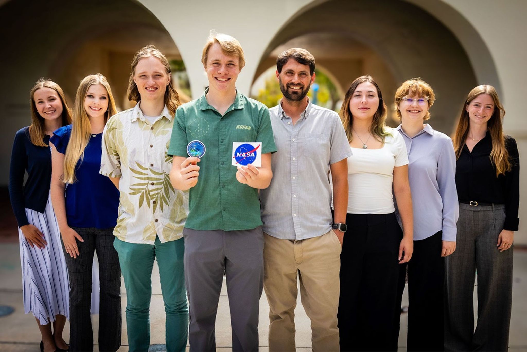 12. Eight people, from SARP West Terrestrial cohort, in business casual attire stand side-by-side smiling for a group portrait in front of a building with stone archways. In the center of the group, a young man holds up a blue mission patch in one hand and a NASA logo sticker in the other.