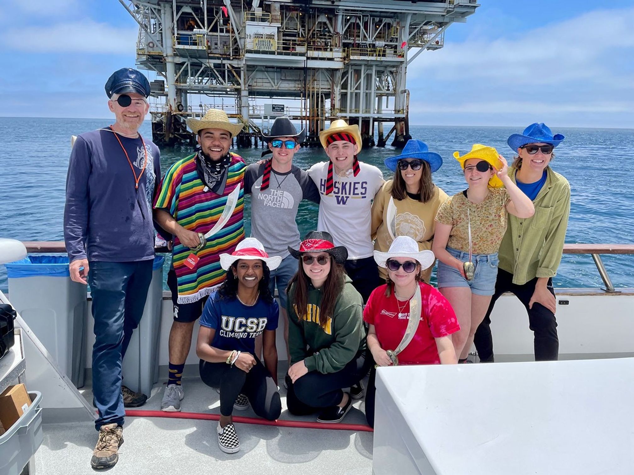 Nine people in a variety of pirate themed dress levels on the edge of a boat, ocean and a research or drilling platform in the background.