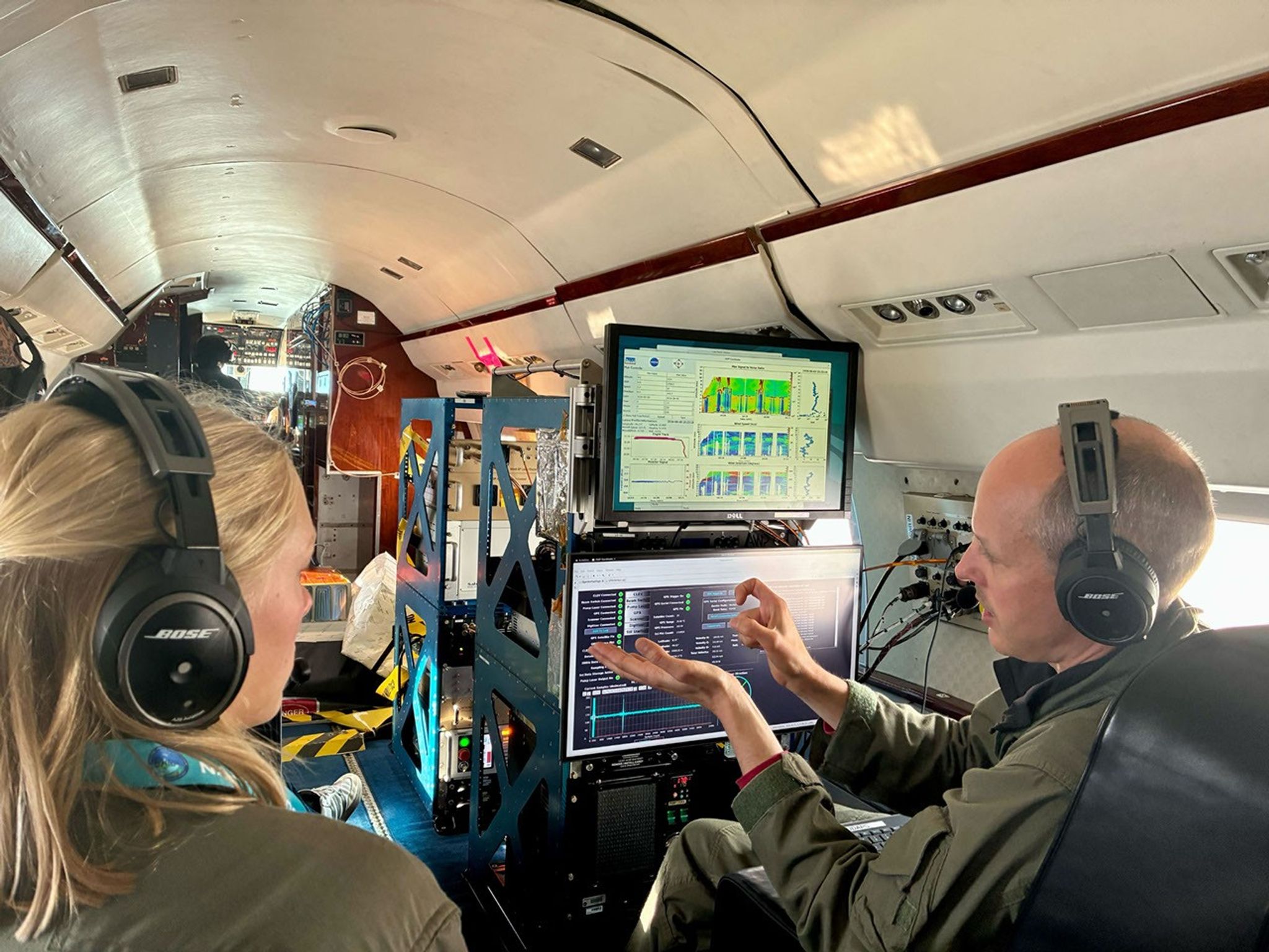 Two people wearing flight suits and aviation headsets discuss scientific data displayed on computer monitors inside a research aircraft cabin during the Student Airborne Research Program.