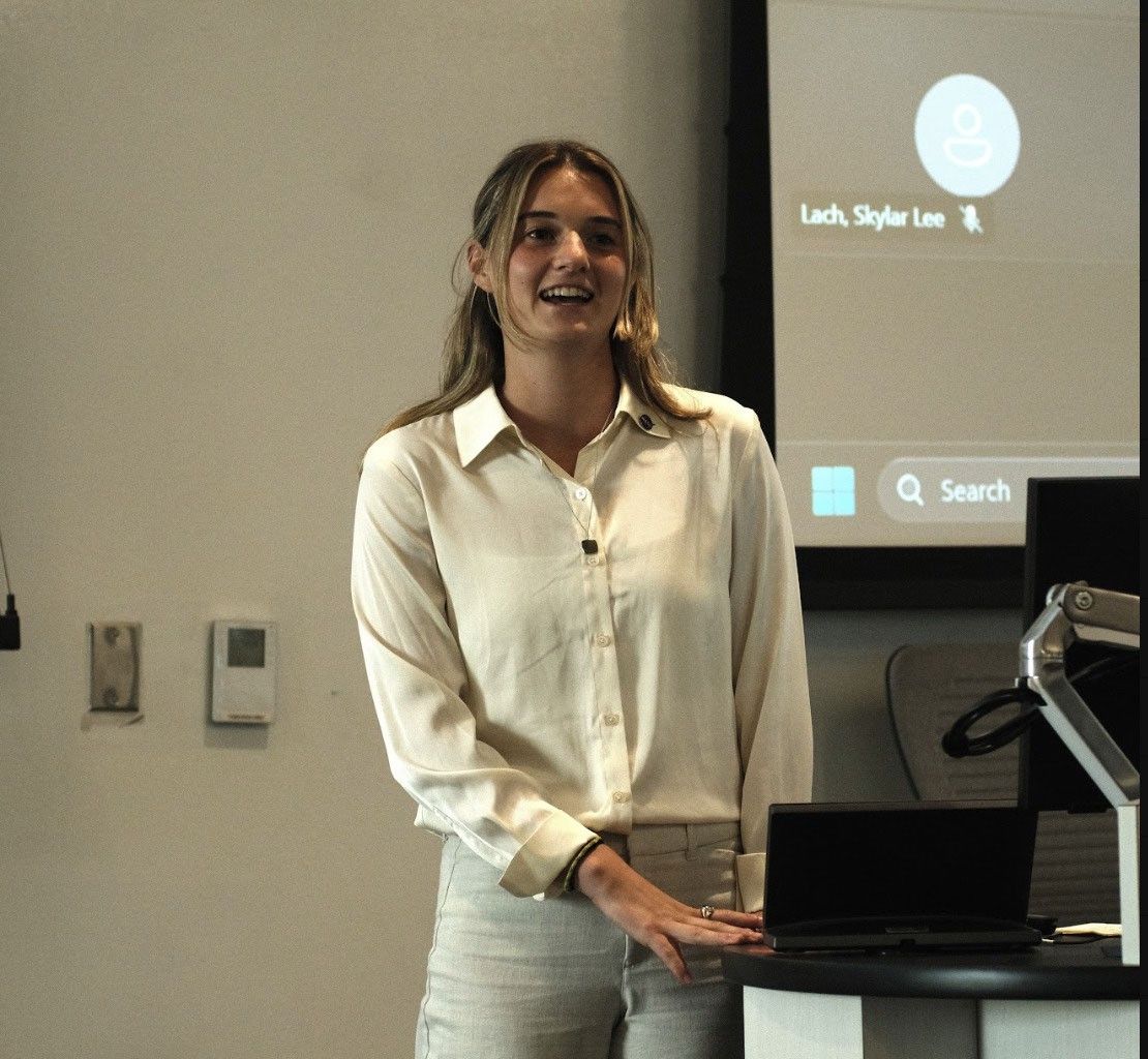 A smiling woman, Catherine Pope, from the Student Airborne Research Program East cohort, in a white button-down shirt gives a presentation at a podium, with a virtual meeting interface projected on a screen behind her.
