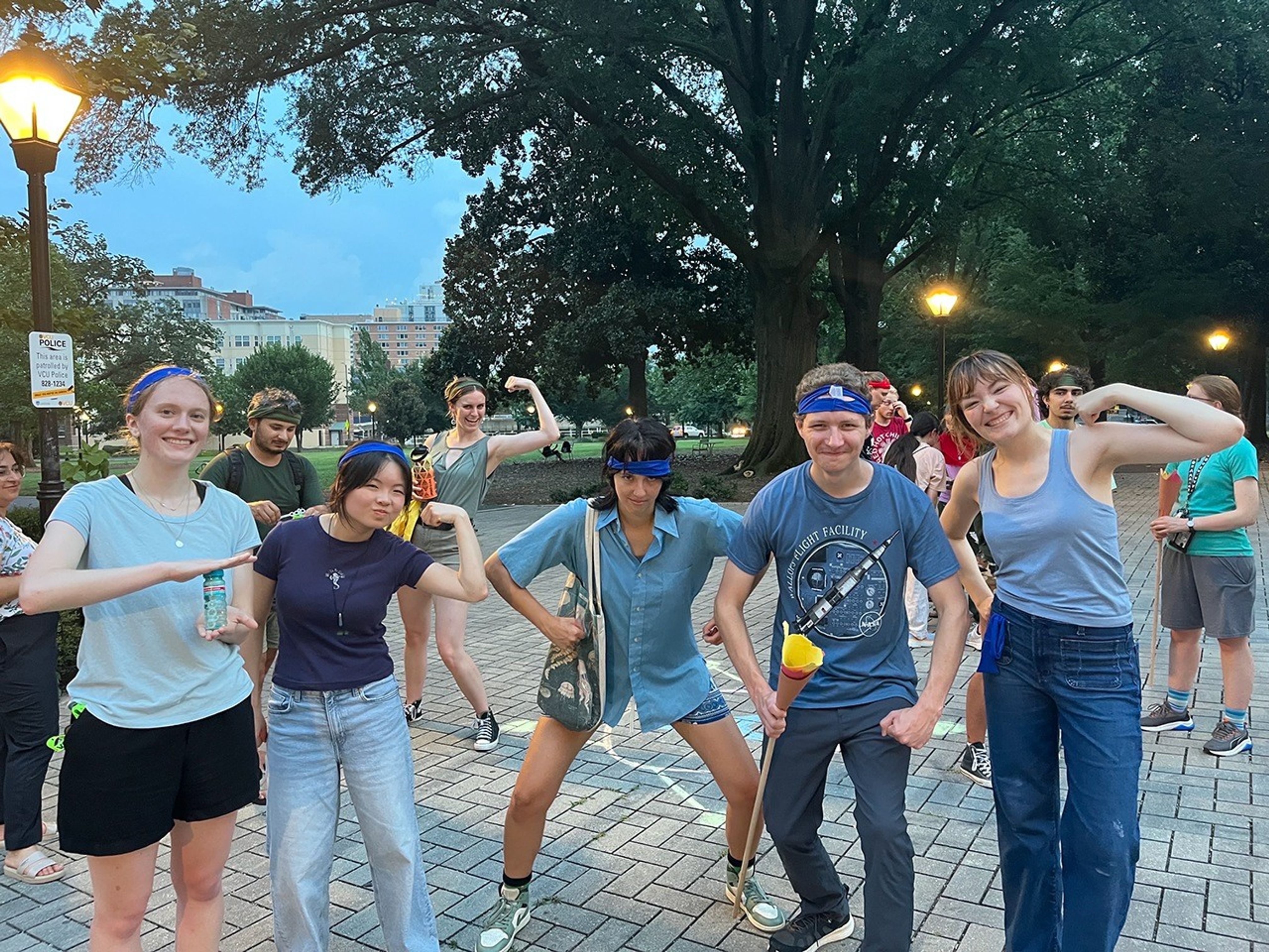 Scattered students with various color headbands pose flexing. They stand on pavers and there are large trees nearby on the right and center of the image. In the background on the left is open sky and tan color buildings.