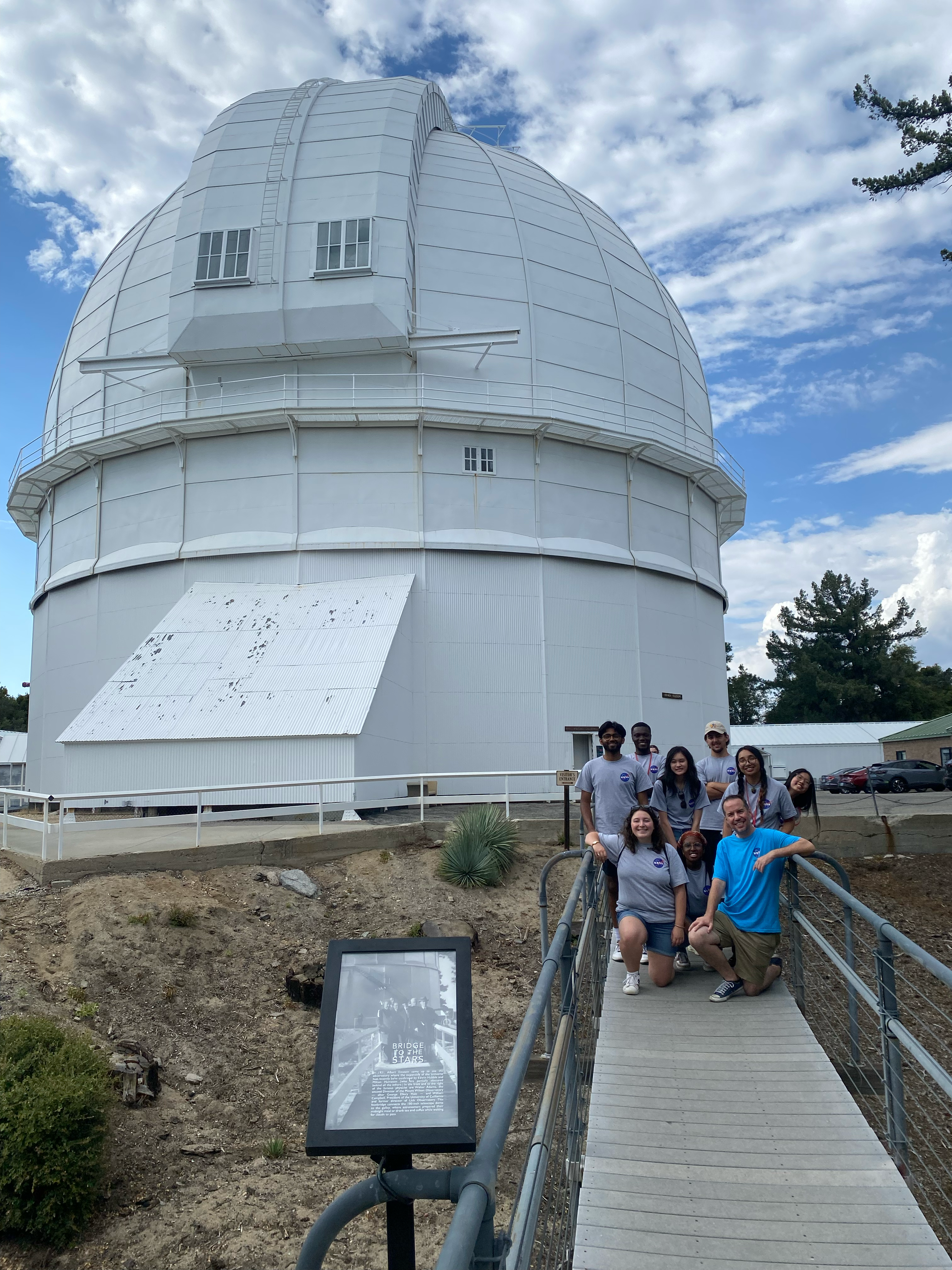 a white domed building with a bridge walkway occupied by 9 people posing for a photo.