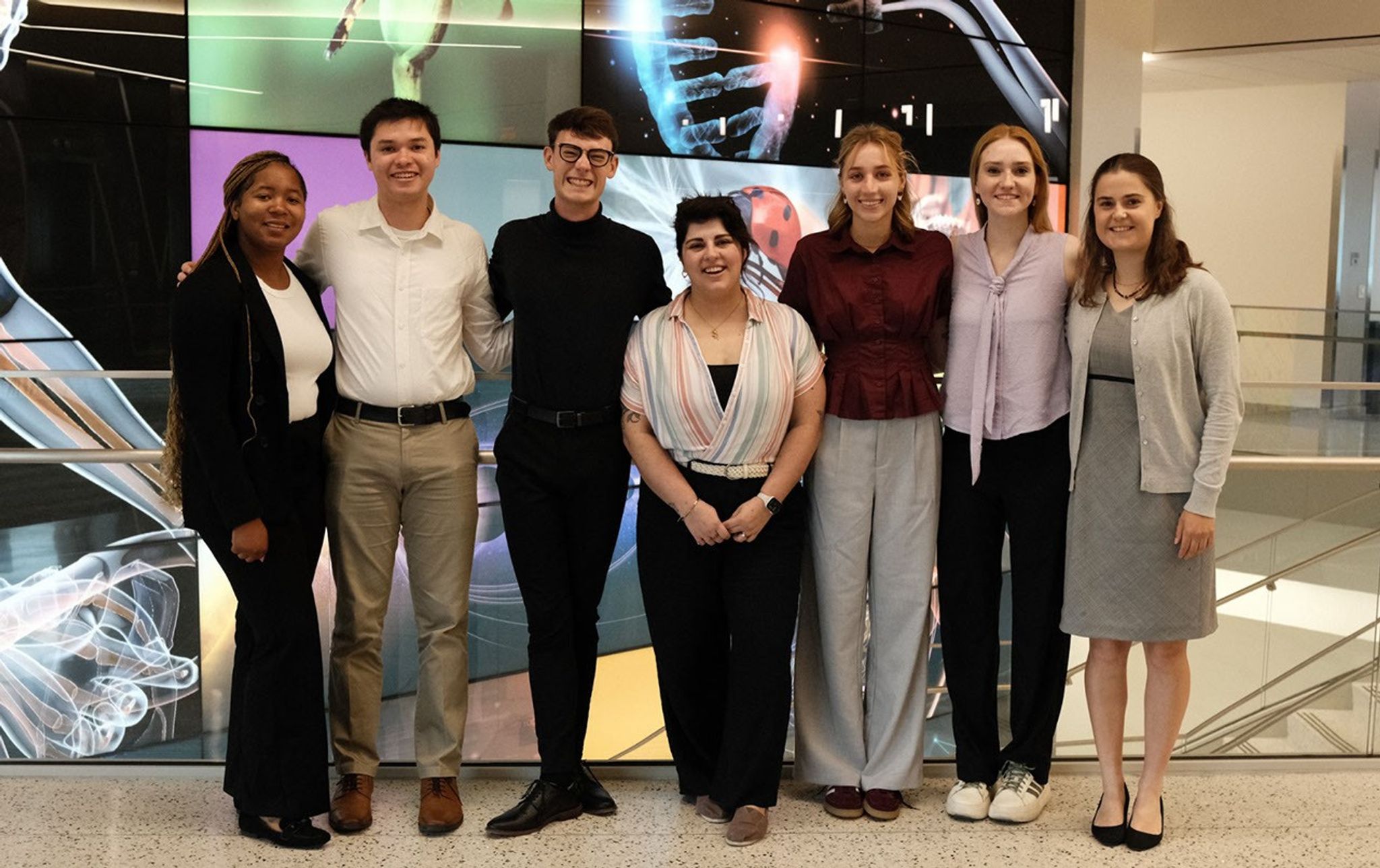 4. Seven professionally dressed young adults, from SARP Oceans East cohort, stand side-by-side for a group portrait in a modern building lobby. Behind them is a vibrant digital wall display featuring various science and biology graphics.