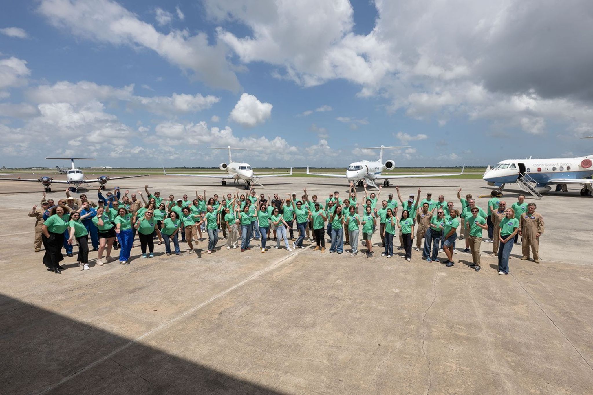 1.	A large group of NASA student airborne research program interns wearing matching bright green t-shirts posing and waving on an airfield tarmac. Several parked airplanes are visible in the background under a sunny, partly cloudy sky.