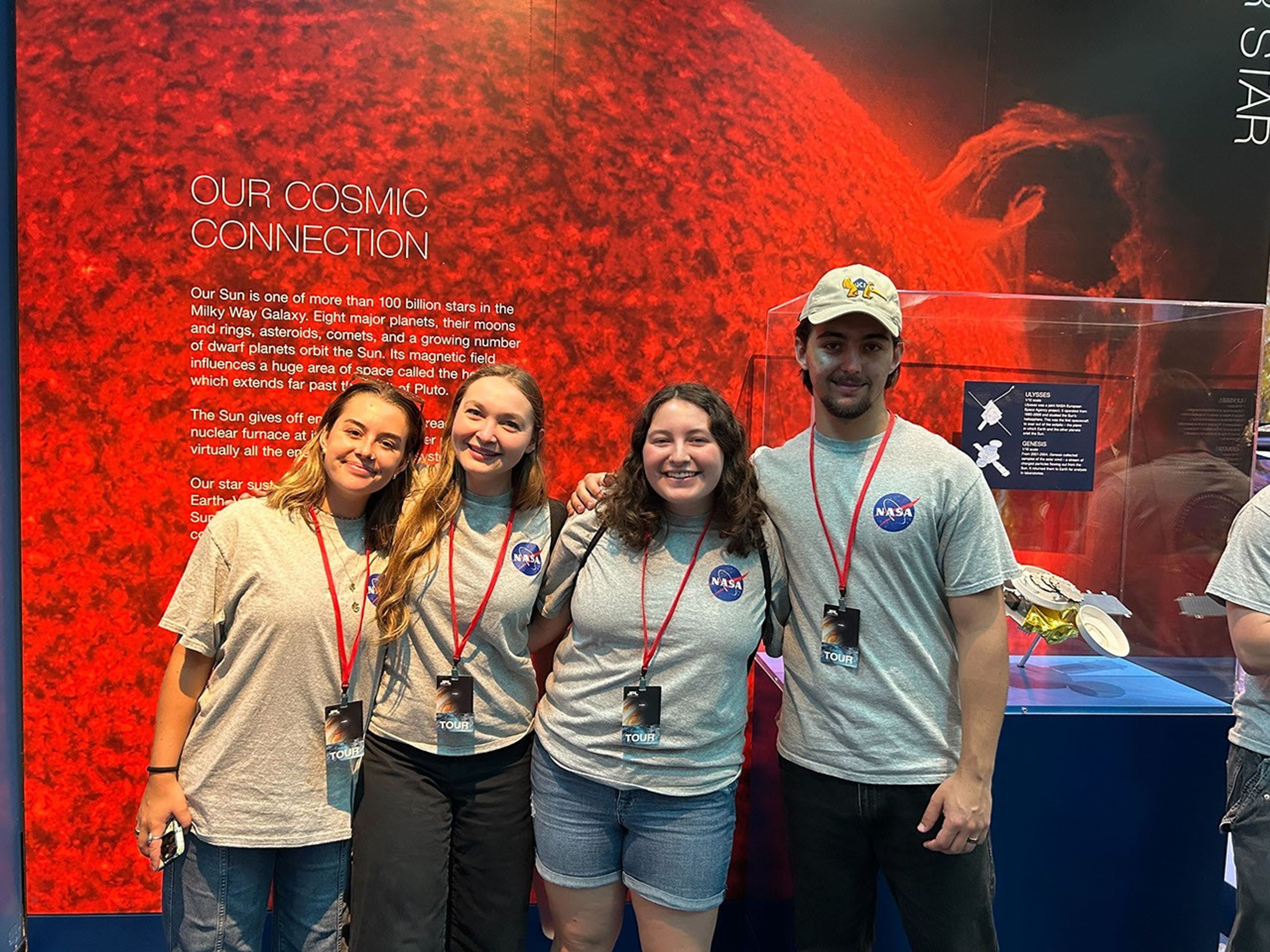 four sarp students in gray t-shirts with the NASA logo stand together for a pose, arms around each other, in front of a vibrant red backdrop and a plexiglass case where something undiscernible is on display
