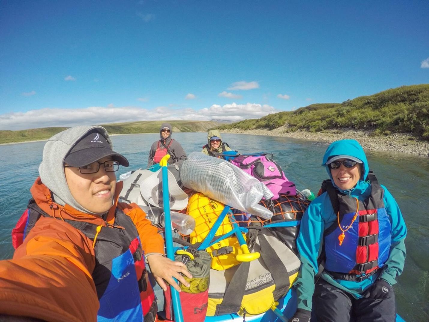 People wearing rain gear and life vests sit on a raft piled high with brightly colored equipment.