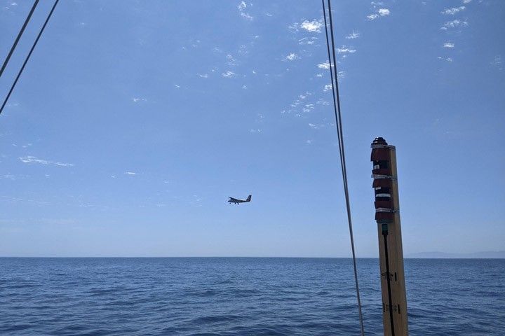 An airplane is visible flying over water as seen from the deck of a sailboat.