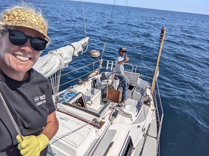 Two people stand on a sailboat surrounded by blue water.