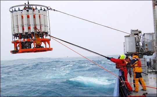 Crew members aboard the R/V Roger Revelle retrieve a CTD rosette from the frigid waters of the Southern Ocean. As the device is lowered into the ocean, electronic instruments measure salinity, temperature, and depth. Each of the white bottles collects seawater at different depths for detailed analysis.