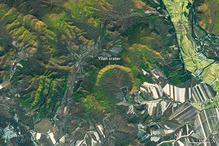 Amid forested landscape that occupies the majority of the image, including the entirety of the upper left to the center of the image, is a crescent shaped ridge labeled the Yilan crater. The Balan river is labeled along the right edge of the image in the upper portion and the surrounding land appears to be segmented into farmed pale green fields. The land along the bottom of the image also consists of many divided strips, though of grays browns and near white colors.