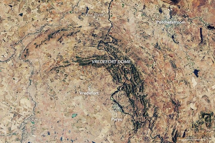 the beige colored landscape is interrupted by an arc of elevated craggy land that is darker in color labeled the Vredefort dome. The Vaal River runs vertically through the this feature as a dark crack on the landscape. Vredefort and the town of Parys are in the lower left from the dome, the latter along the river. Another town is visible in the top right labeled potchefstroom
