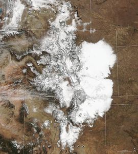 The satellite image focuses on Colorado and surrounding states, the state lines are superimposed on the landscape along with state names. The landscape is bright white where snow covers the ground, and elsewhere is a deep brown color.