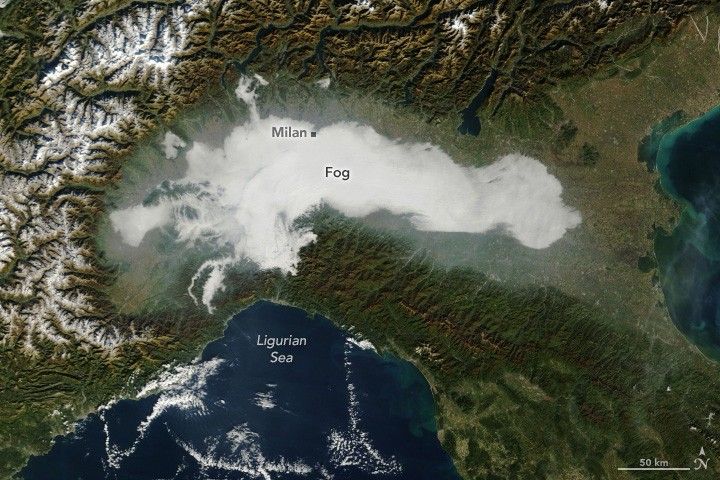 The satellite image shows Northern Italy valley with dense fog coverage. At the top of the image are the snow covered alps, at the bottom the Ligurian Sea. Milan is labeled under the northern edge of the fog.