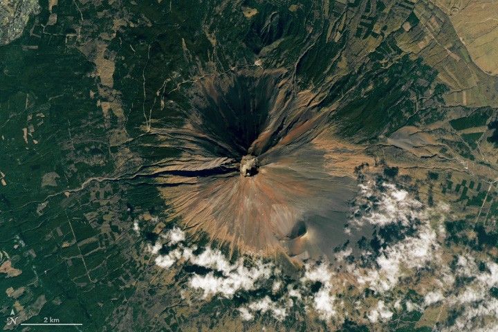 A top down view of Mount Fuji. In the exact center is the crater which matches the color of the surrounding circular field of slopes, primarily light brown. To the lower right the color takes on a more faded black look. Sparse clouds occupy the lower right of the image beyond the volcanic slopes. The remainder of the image gives way to deep green foliage and lighter patches, possibly farmed fields, primarily on the upper right portion of the image.