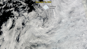 Visible through a layer of thin clouds, a patch of  somewhat square shaped ice is visible on the dark ocean waters. It is labeled Iceberg A-23A just under the date label of December 16, 2024