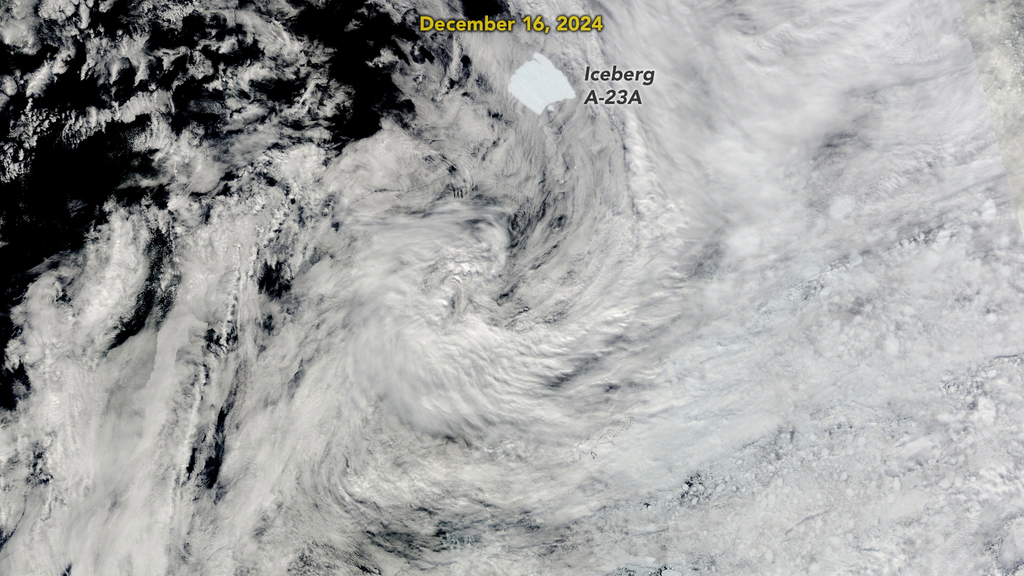 Visible through a layer of thin clouds, a patch of  somewhat square shaped ice is visible on the dark ocean waters. It is labeled Iceberg A-23A just under the date label of December 16, 2024
