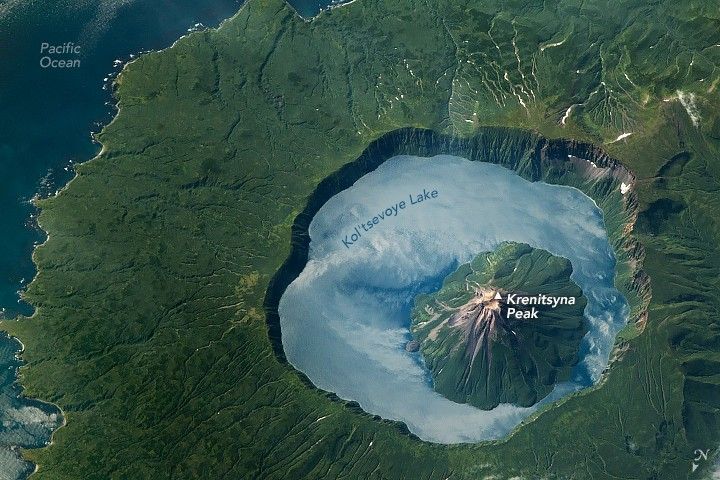 Krenitsyna peak features in the lower right of the image surrounded by Kol’tsevoye Lake surrounded by the cliffs of a caldera. The land gradually slopes down from the peak of the caldera, covered in dark green foliage before meeting the Pacific Ocean along the left side of the image