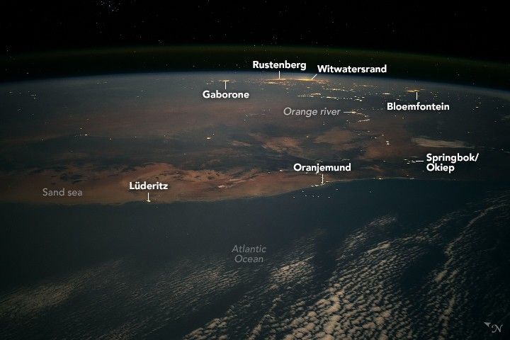 Photo of Earth from the ISS shows the Atlantic Ocean in the foreground with some sporadic cloud coverage. Along the coast is the town of Oranjemund, Lüderitz and the Sand sea. Slightly inland is Springbok/Okiep. Near the end of visible Earth is Gaborone, Rustenberg, Witwatersrand, and Bloemfontein. Each of these settlements appear illuminated by human-made lighting. The Orange river is also illuminated by the peoples who live along the river.