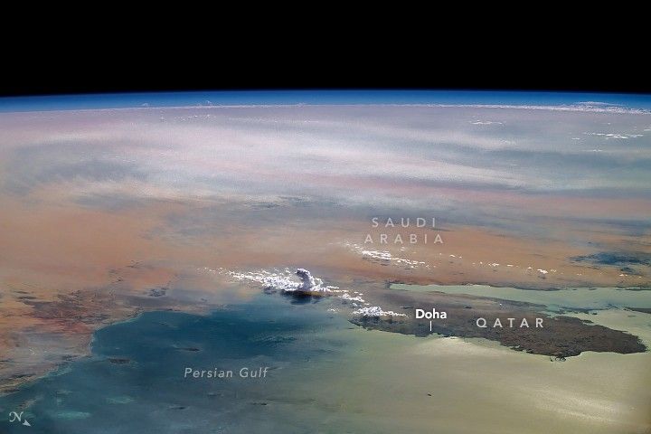 In the foreground of the earth image is the Qatar peninsula on the Persian Gulf. To the left of Doha is a single towering cloud  amid other clouds. The surrounding land is a shade of brown, further in the background of the image the land is overcast in gray before the blue haze of atmosphere gives way to the darkness of space.