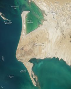 The Cheleken Peninsula, home to the town of Hazar, centers the image, with mainland to the right and Caspian sea left. The land is all a light beige color with dried lakes causing elongated swirling patterns of slightly darker land and dunes giving the land a textured look above that. Peninsula Kafaldja extends upward off of the Cheleken and the Dervish downward. Both peninsulas are extremely narrow . The waters at the top of the image, in the Türkmenbaşy gulf are a lighter shade, even a vibrant green near the peninsula. The Türkmenbaşy peninsula appears disconnected from land at the far left edge of the image. An Oil rig appears as a small dot in the middle of the shown portion of the Caspian Sea.