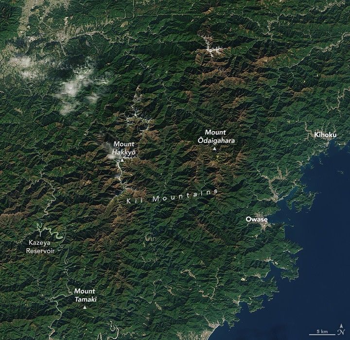Two of the Kii Mountains, Hakkyō and Ōdaigahara, show peaks having a lustrous reddish brown color. Human settlements are clearly visible in the valley at the top left corner, Kazeya Reservoir is shown at the lower left, but above Mount Tamaki. Ocean occupies the lower right along with the coastal towns of Owase and further up the coast Kihoku.