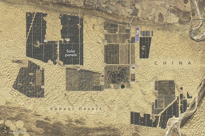 There are numerous fields of solar panels, dark gray in color divided by lines and dots of soil in the mottled beige landscape of the Kubuqi Desert in China. Toward the bottom of the image the land takes on a darker shade and at the top a road runs the length of the image, above which seems to be distinct fields, also primarily beige. At the bottom left are some perfect circles of land, seemingly smoother than the surrounding terrain and mostly lighter in color than their surrounds. Two separate clusters are all close to the point of potentially overlapping.