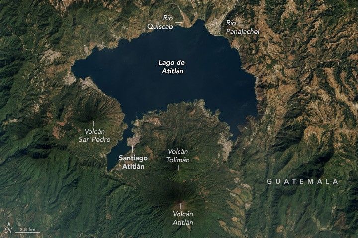 Lago de Atitlán, dark blue, is nestled into the landscape, with human settlements along the coast and Volcanos, covered in deep green foliage, around the lower coastline. Volcán Atitlán is labeled, near the bottom center of the image. Just above that and closer to the lake is Volcán Tolimán which base spreads increasingly gradually to the lake shore.  On the left edge of Volcán Tolimán the municipality of  Santiago Atitlán. A narrow strip of the lake separates this settlement from Volcán San Pedro.  At the top of the lake are the river deltas of Rio Panajachel and Rio Quiscab