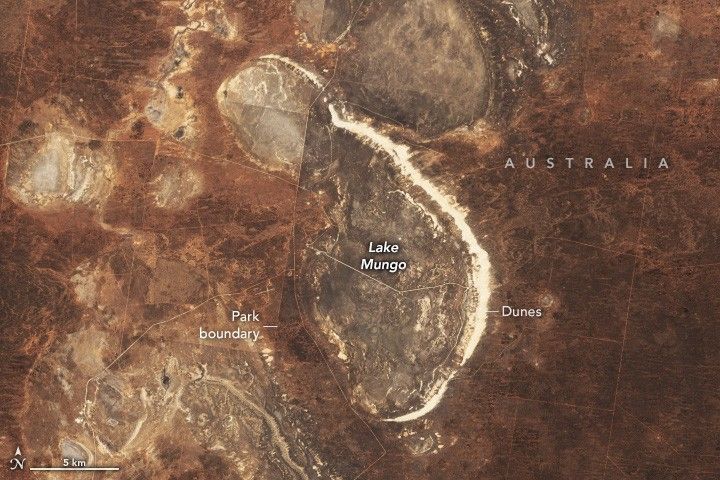 A rust brown colored landscape is interrupted by a lighter brown Lake Mungo in the center of the image. Along the right edge of Lake Mungo are bright white Dunes. To the left of most of Lake Mungo is a Park boundary line that seems to separate a lighter shade of landscape on the left from a darker one on the right.
