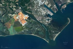 Along the left bank of the Rhône River, orange to white rectangular salt pans are interwoven with coastal marsh along the Gulf of Lion at the bottom of the image. Additional gridded land along the coast, potentially also salt pans, vary in color from burnt orange, to seafoam and verdant green stand apart. To the right of the Rhône River is Port-Saint-Louis-du-Rhône. The shipping port and human settlement area  landscape appears streaked with gray and white around the gray concretes and brown roofed buildings.