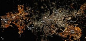 The dark image is filled with city lights. 6 October city shows up in amber lights on the left. A dark patch between that and Giza is labeled pyramids of Giza. A dark line, the Nile River separates the area of white light, Giza Left and Cairo right, in the center of the image. In the bottom right is more amber lighting and it it labeled New Cairo. The airport is just above New Cairo.
